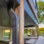 Close-up of a modern house exterior showing a metal downspout attached to a wooden post. Outdoor kitchen and patio furniture are visible, with trees and a lake in the background. The scene is bathed in natural light.
