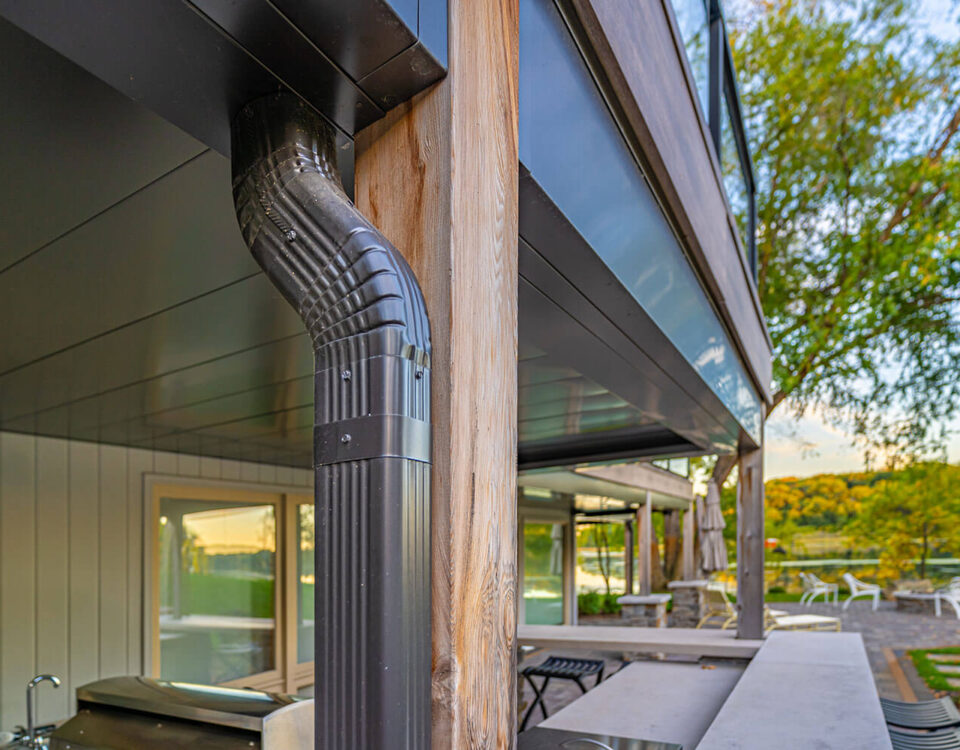 Close-up of a modern house exterior showing a metal downspout attached to a wooden post. Outdoor kitchen and patio furniture are visible, with trees and a lake in the background. The scene is bathed in natural light.