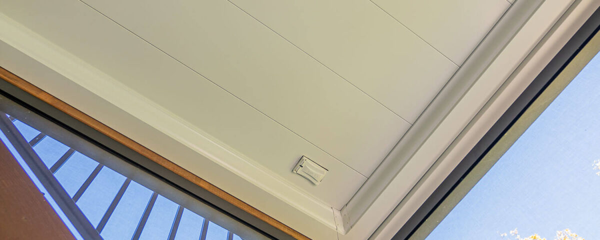 View looking up at a modern, white paneled ceiling with a small circular light and a smoke detector. Part of a glass door, metal railing, and green tree branches are visible in the corner.