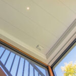 View looking up at a modern, white paneled ceiling with a small circular light and a smoke detector. Part of a glass door, metal railing, and green tree branches are visible in the corner.