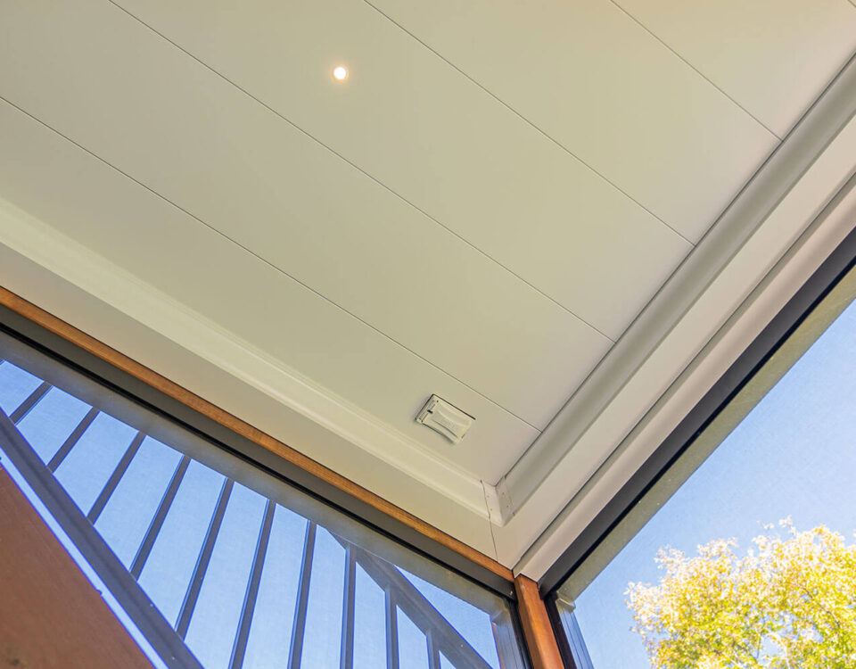 View looking up at a modern, white paneled ceiling with a small circular light and a smoke detector. Part of a glass door, metal railing, and green tree branches are visible in the corner.