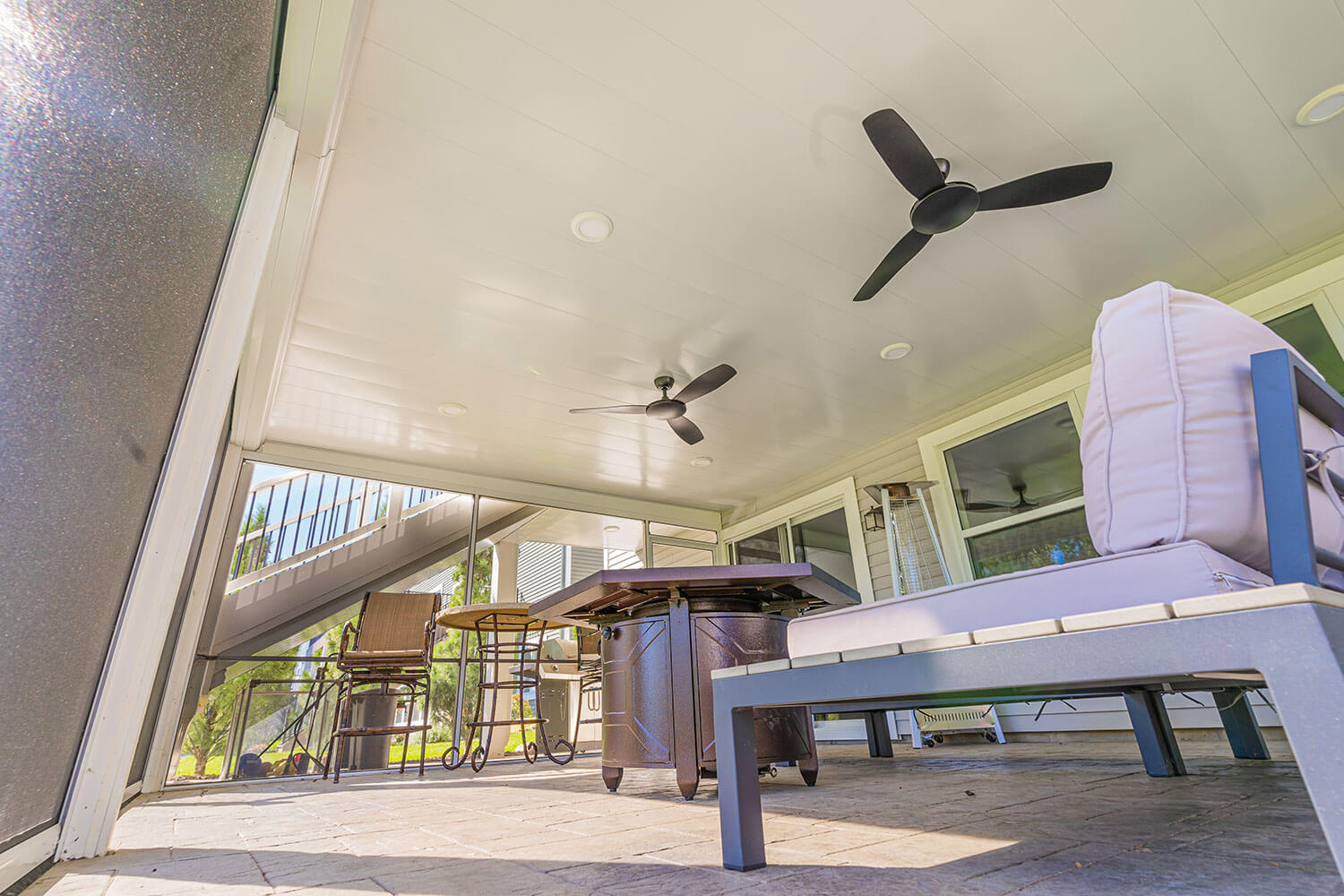 A covered patio with ceiling fans, cushioned seating, a table, and a grill. The area is well-lit with natural light coming from large windows and a screen, overlooking a staircase and green lawn outside.