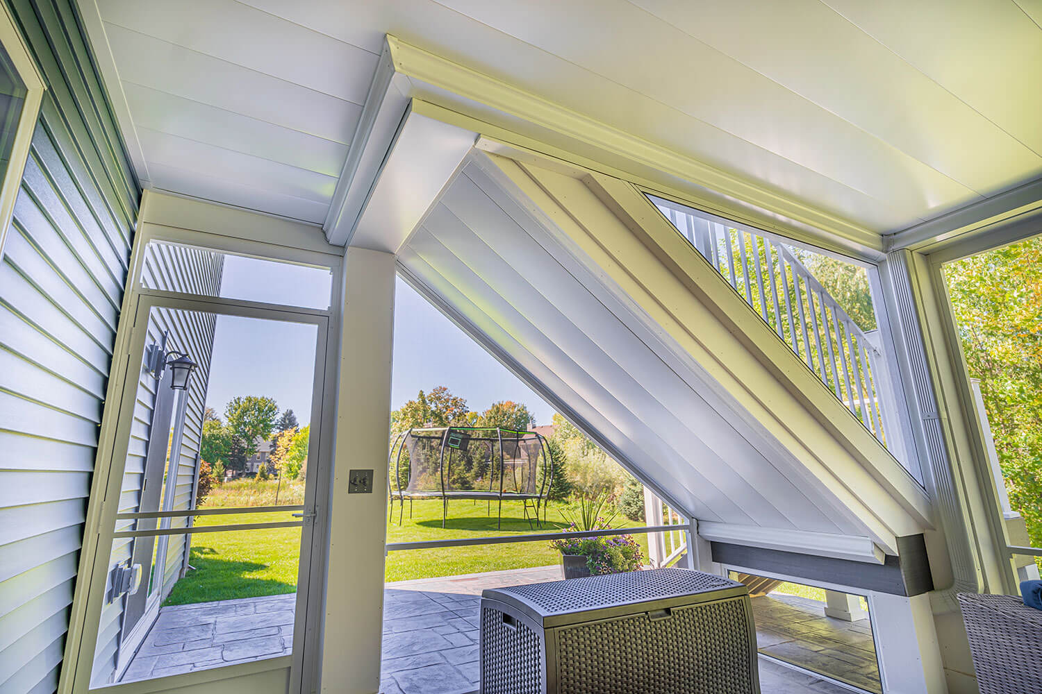 A screened-in porch with a slanted, partially open louvered roof, letting sunlight in. The view shows a green backyard with trees and a trampoline. Patio furniture sits on a stone floor.
