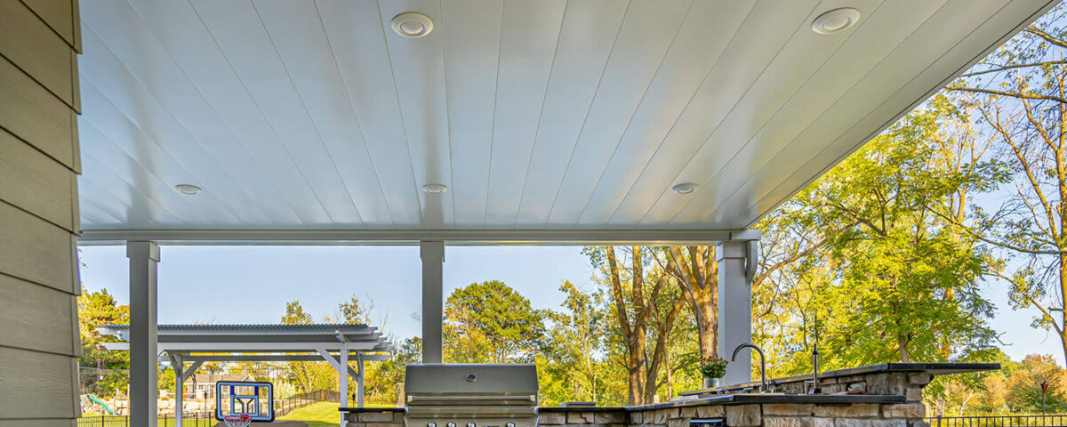 Covered outdoor patio with a built-in stone grill and sink, ceiling lights overhead, and a view of a pool area and basketball hoop in a landscaped backyard with trees.