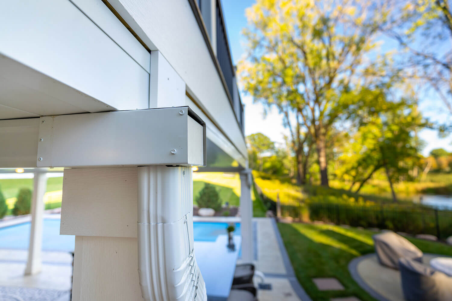 Close-up of a white gutter and downspout system attached to a deck post, with a backyard pool, patio area, green lawn, and trees blurred in the background on a sunny day.