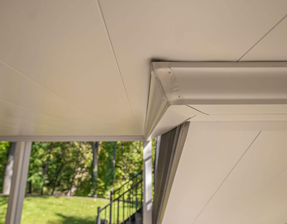 Close-up view of a clean, modern white ceiling with angled panels and metal trim, part of a covered outdoor patio; green trees and a black staircase are visible in the background.
