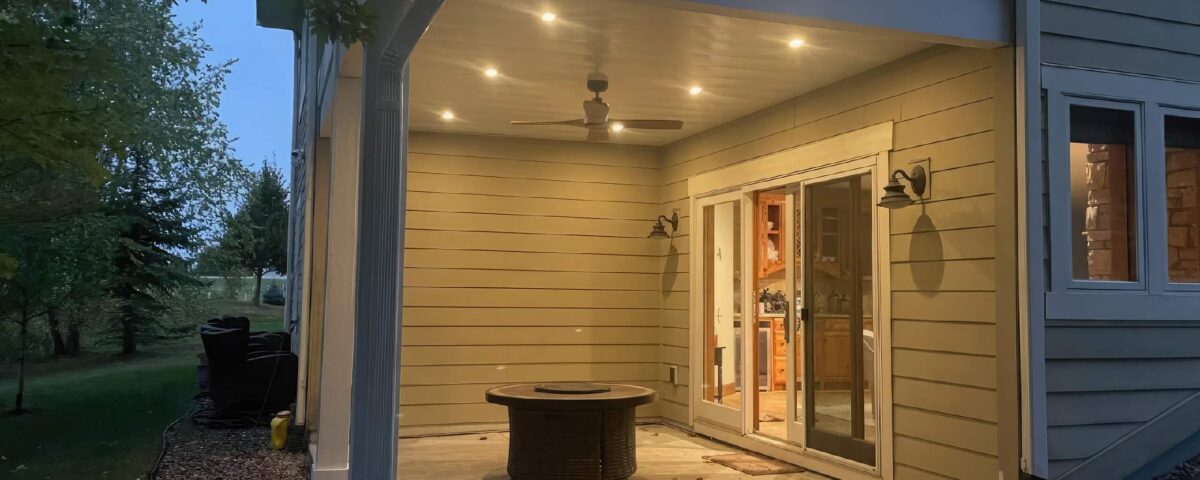 A covered patio with recessed lighting and a ceiling fan, featuring a round fire pit on a concrete floor. Glass doors lead inside the house, and the patio is surrounded by landscaping rocks and a grassy yard.