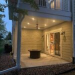 A covered patio with recessed lighting and a ceiling fan, featuring a round fire pit on a concrete floor. Glass doors lead inside the house, and the patio is surrounded by landscaping rocks and a grassy yard.