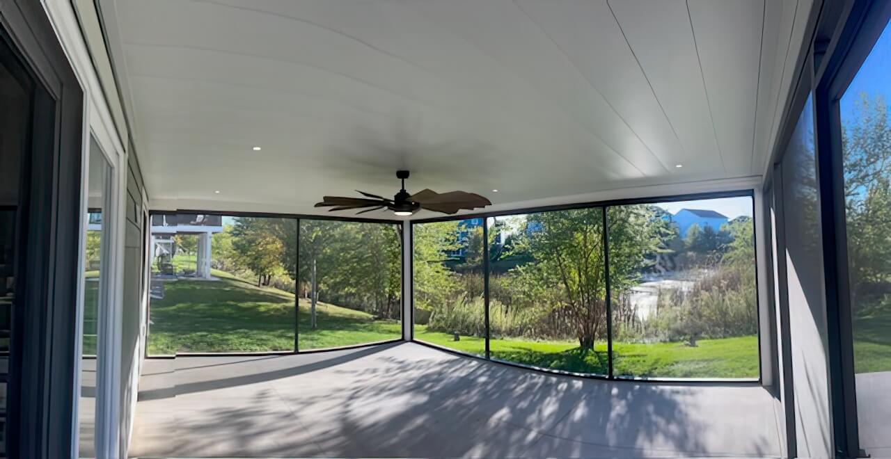 A spacious screened-in porch with a ceiling fan overlooks a green yard with trees and a pond. Sunlight filters through large floor-to-ceiling windows, creating shadows on the gray floor.