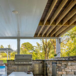 Outdoor covered underdeck area with a built-in stone kitchen, stainless steel grill, and sink. Exposed wood beams overhead; pool with a basketball hoop and lounge chairs visible in the background, surrounded by trees.