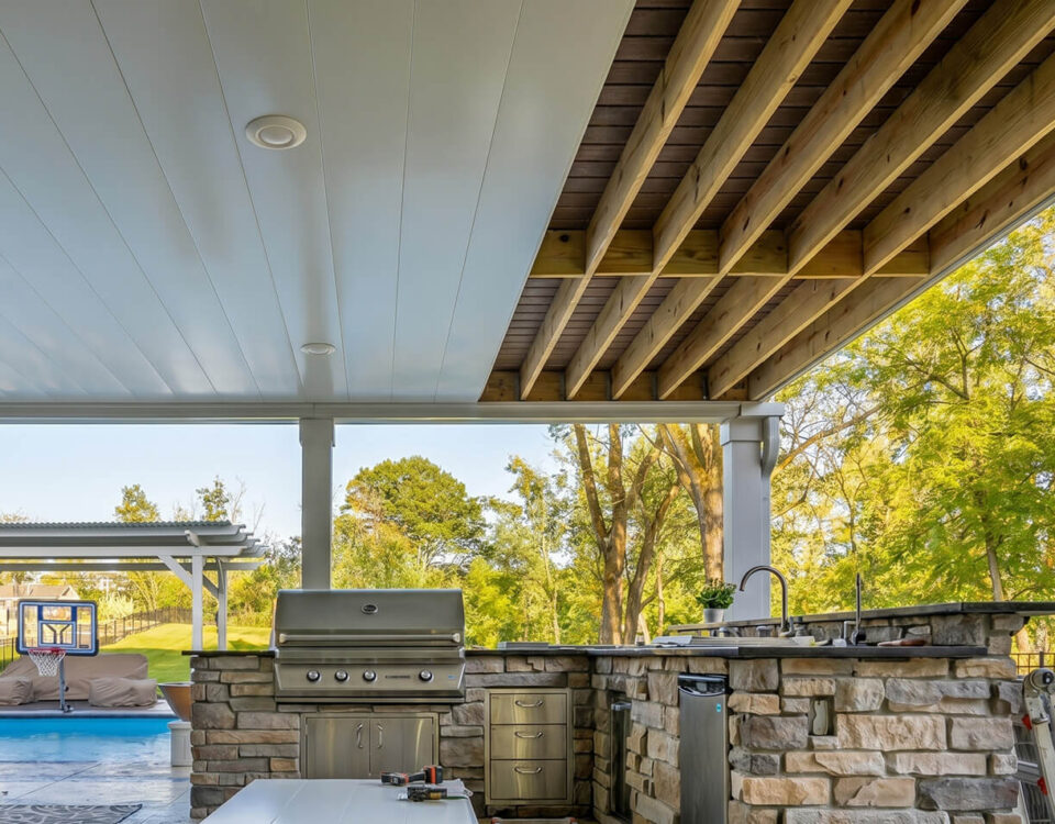 Outdoor covered underdeck area with a built-in stone kitchen, stainless steel grill, and sink. Exposed wood beams overhead; pool with a basketball hoop and lounge chairs visible in the background, surrounded by trees.