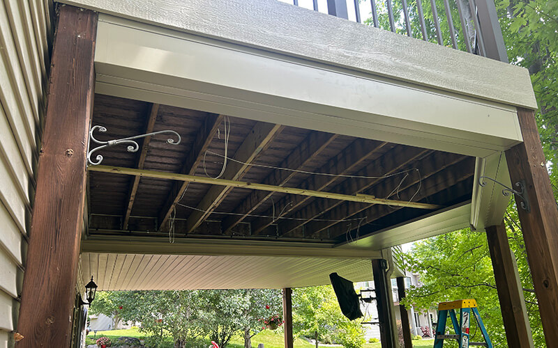 A Magnolia underdeck being installed in the surface style. View of the underside of an elevated deck with exposed beams and wires, supported by wooden posts, with a partial view of a backyard, greenery, and a blue ladder in the background.