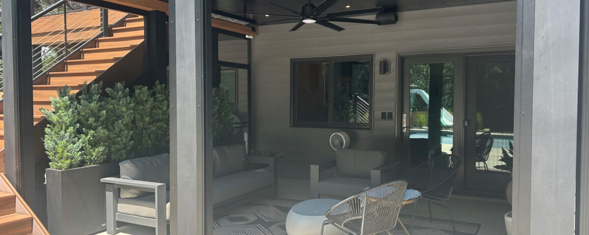 Underdeck covered patio with gray furniture, a ceiling fan, potted plants, and a patterned rug. Wooden stairs lead up on the left. Glass doors and windows reflect sunlight in the background. Modern, shaded seating area.