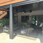 Underdeck covered patio with gray furniture, a ceiling fan, potted plants, and a patterned rug. Wooden stairs lead up on the left. Glass doors and windows reflect sunlight in the background. Modern, shaded seating area.