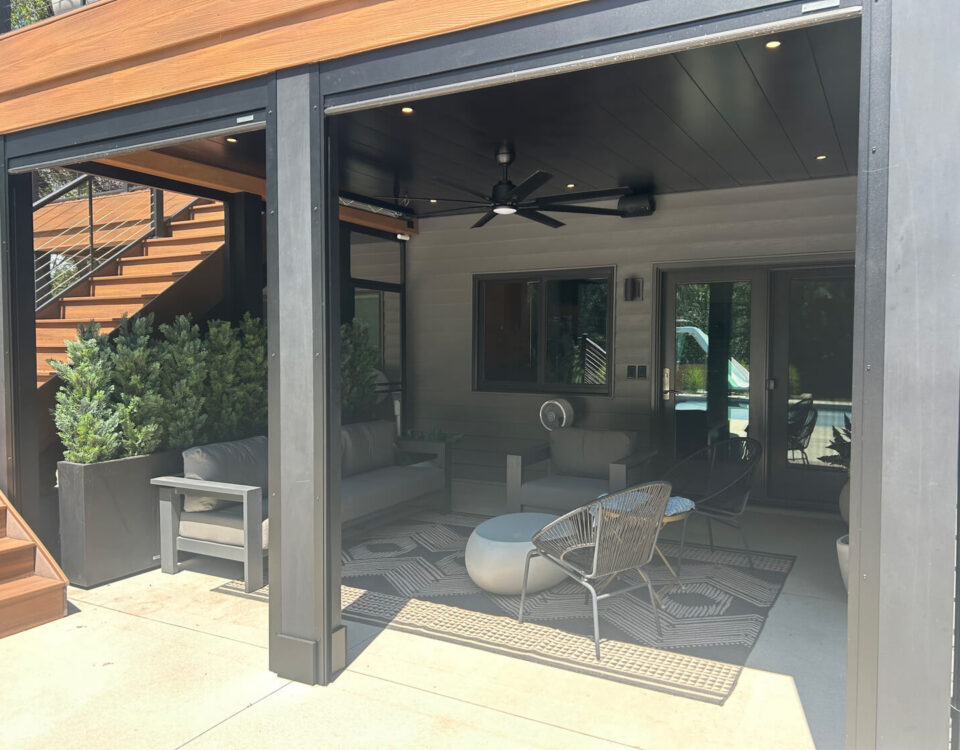Underdeck covered patio with gray furniture, a ceiling fan, potted plants, and a patterned rug. Wooden stairs lead up on the left. Glass doors and windows reflect sunlight in the background. Modern, shaded seating area.